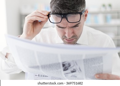 Businessman In The Office, Reading Bad Breaking News On A Financial Newspaper, He Is Shocked And Adjusting His Glasses