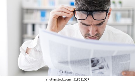 Businessman In The Office, Reading Bad Breaking News On A Financial Newspaper, He Is Shocked And Adjusting His Glasses