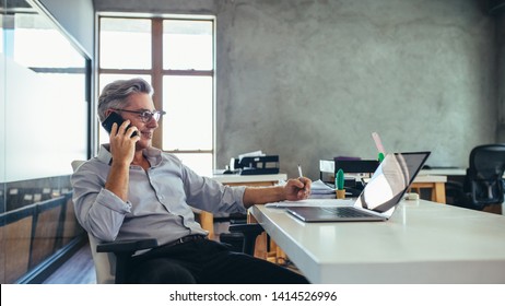 Businessman on the phone sitting at the laptop in his office. Male business professional in office talking on cell phone. - Shutterstock ID 1414526996