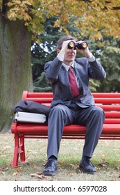 Businessman On A Red Park Bench Looking Through Binoculars