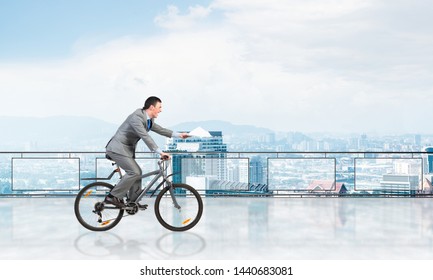 Businessman with paper documents in hand on bike. Deadline for paperwork. Corporate employee in grey business suit riding bicycle on balcony. Cyclist on terrace with office buildings view.