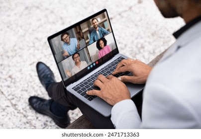 A businessman is participating in a video conference call on his laptop while sitting outdoors in an urban environment. Five colleagues appear on the screen in a professional, virtual meeting. - Powered by Shutterstock