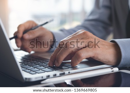 Businessman with pencil in hand working on laptop computer keyboard in modern office, online job concept. Man typing on laptop, searching internet with digital tablet on office desk, online working