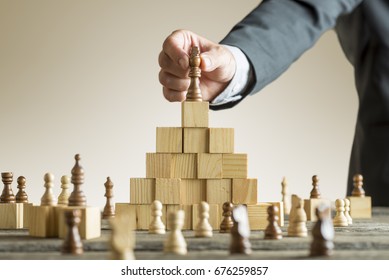 Businessman placing a chess piece on a pyramid of wooden building blocks in a concept of success and achievement in a close up view of his arm. - Powered by Shutterstock