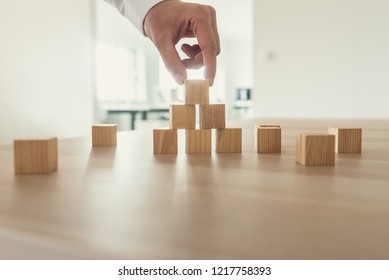 Businessman Placing Wooden Cubes In A Pyramid Shape On Office Desk In His Workplace.