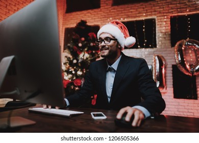 Businessman Playing On Computer On New Year Eve. Christmas Tree In Office. Laptop On Desk. Business Concept. Man In Black Suit. Celebrating Of New Year. Using Digital Device. Red Cap.