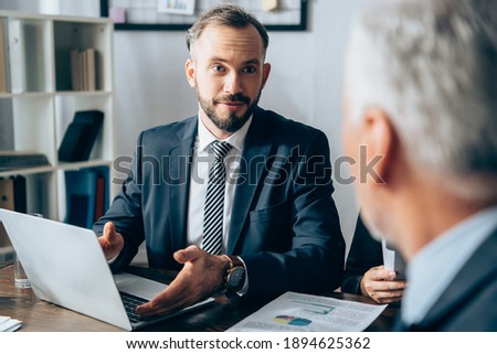 Businessman pointing at laptop near papers and investor on blurred foreground in office