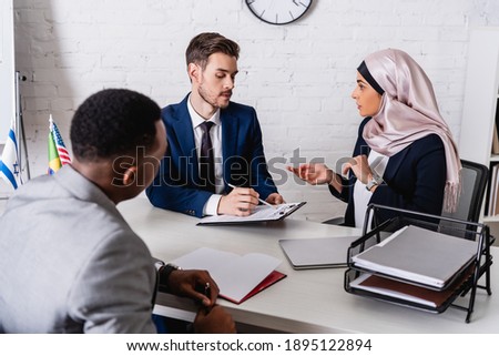 businessman pointing with pen at contract near arabian businesswoman and interpreter, blurred foreground
