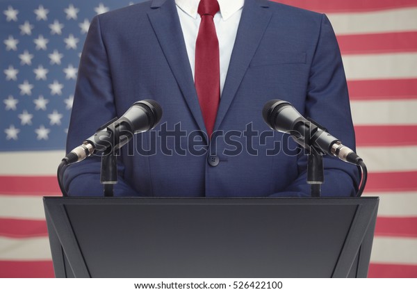 Businessman or politician making speech from\
behind the pulpit with USA flag on\
background
