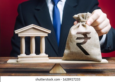 Businessman Puts A Indian Rupee Money Bag On Scales Opposite To Government Building, Bank, University. Lobbying Interests. Taxes Payment. Deposits, Loans. Financial Support For Reforms. State Debt