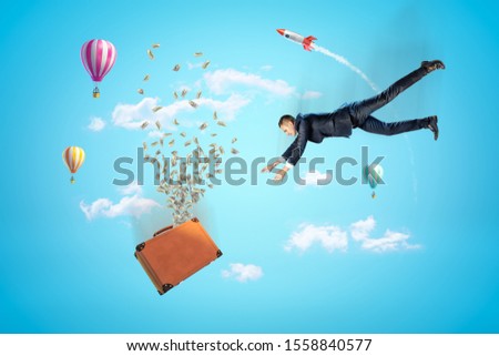 Businessman reaching to brown suitcase with dollars flying out, hot air balloons and silver red space rocket in the air on blue background. People and objects. Banking and finance. Holidays and