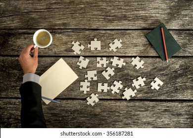 Businessman reaching for a cup of coffee on his rustic wooden desk as he searches for solution to a problem  with puzzle pieces scattered on his desk next to a blank paper and notepad with a pen. - Powered by Shutterstock