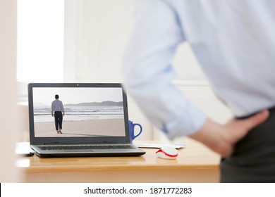 Businessman Relaxing At His Workplace At Home While Looking Wistful On A Laptop With Businessman Walking On A Beach - Same Model On The Screen And In The Foreground