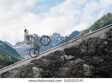 Businessman riding uphill by bike. Nature landscape with copy space. Man in business suit riding bicycle on mountain road. Cyclist popped wheelie on background of blue sky. Healthy lifestyle