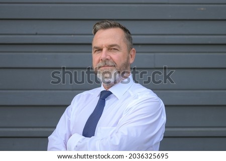 Businessman scrutinising the camera with a serious assessing look as he stands with folded arms in front of a grey exterior wall
