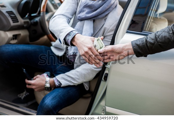 Businessman sitting in car with open door gives\
twenty dollars to another\
man