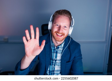 Businessman sitting at his desk wearing headset having online meeting while working late in an office, smiling and waving towards the camera - Shutterstock ID 2255602087