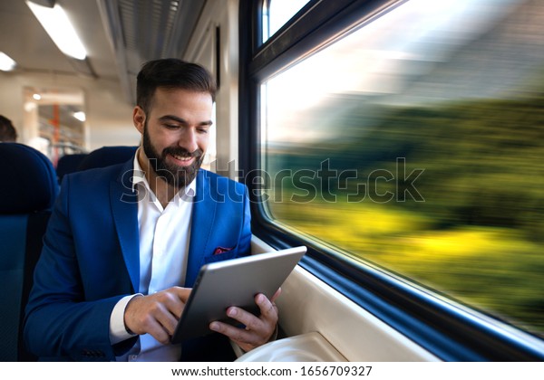 Businessman sitting next to window reading news\
and surfing internet on his tablet while traveling in comfortable\
high speed train.