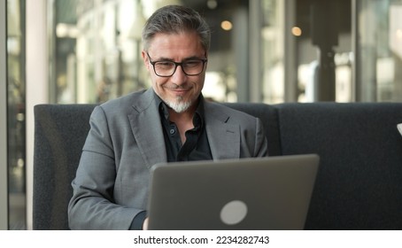 Businessman sitting on couch working with laptop computer in office lobby. Portrait of happy middle aged man in business casual, smiling. - Powered by Shutterstock