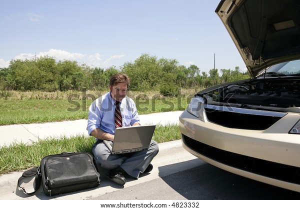 A businessman sitting on the curb working beside his\
broken down car.