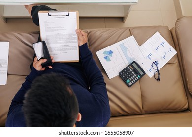 Businessman Sitting On Sofa With Financial Charts And Reading Partnership Agreement, View From Above