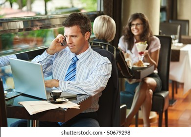 Businessman sitting at table in cafe using laptop computer, talking on mobile. - Powered by Shutterstock