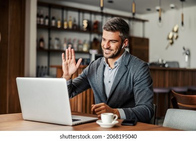 A businessman is sitting in working friendly cafe with earphones in his ears and having a conference call over a laptop. The man is waving to the colleagues. A businessman having a video call - Powered by Shutterstock