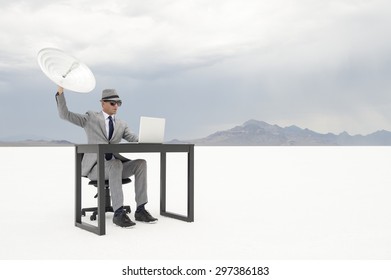 Businessman Sitting Working At His Outdoor Office Desk Trying To Communicate Holding Up A Satellite Dish In Stark White Desert Landscape