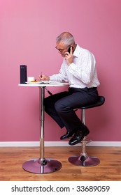 Businessman In Smart Casual Attire Sat On A Bar Stool In A Cafe Working Through His Lunch Break.