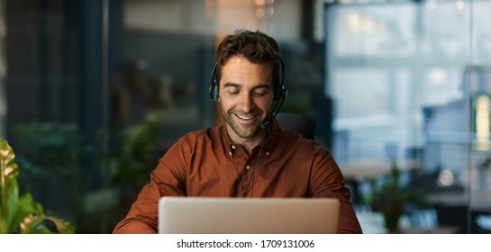 Businessman smiling and talking with a client over a headset while working alone at his desk in a dark office after hours