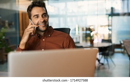 Businessman Smiling And Talking On His Cellphone While Working On A Laptop At His Desk In An Office After Hours