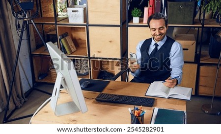 Businessman smiling while using his phone, scrolling through social media instead of focusing on work laid out in front of him. Concept of business, people and daily work challenges. Ad.
