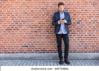 Businessman Sms Texting Phone App In City Street On Brick Wall Background. Business Man Holding Smartphone In Smart Casual Wear Standing. Urban Young Professional Lifestyle.