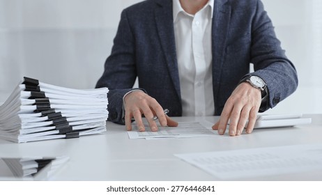 Businessman sorting through stacks of documents on a white desk beside a tall pile of bound papers, symbolizing meticulous financial record keeping and bureaucracy. Audit and taxes