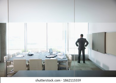 A Businessman Standing In A Conference Room With Hands On His Hips While Looking Outside The Glass Window