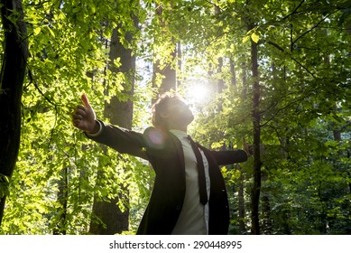 Businessman standing with his arms outspread showing thumbs up sign celebrating business success in woodland with fresh green leaves on the trees backlit by the rays of the sun, low angle view.