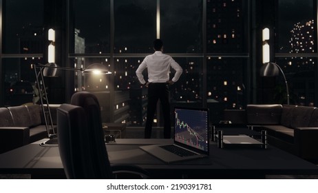 Businessman Standing In His Office Working Late At Night, Looking Out Of The Window As It Rains. Corporate Office With Realtime Stock Trading Charts On Laptop Computer Showing Recession Bear Market.