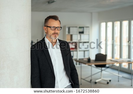 Businessman standing leaning against an office wall staring aside with a serious expression as he plans his strategy or reaches a decision