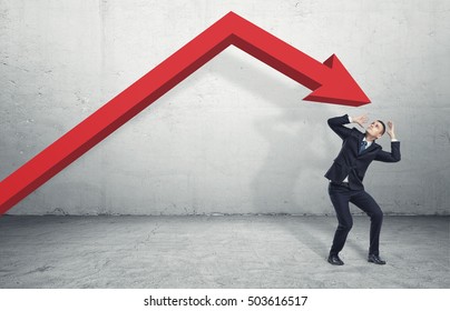 Businessman standing on the floor in a desparate pose under the giant red arrow aiming down at him, on the grey background. Business and management. Problems and failures. Dealing with crisis.