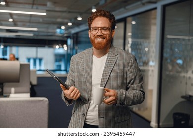Businessman standing on office background with coffee and tablet and looks camera with smile - Shutterstock ID 2395459447