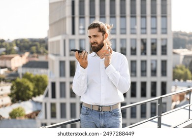 Businessman standing on rooftop using smartphone with urban background. Man in white shirt and jeans speaking into mobile phone, city buildings in the background. Professional communicating outdoors. - Powered by Shutterstock