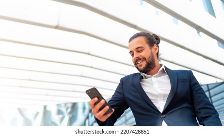 Businessman Standing Outdoor Use Smartphone Looking Device Screen And Smile. Handsome Hispanic Male Business Person In Business Suit Holding Smartphone In Hand. Modern Business Person Low Angle View