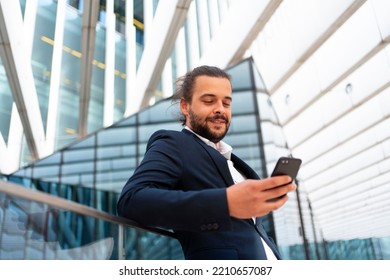 Businessman Standing Outdoor Use Smartphone Looking Device Screen And Smile. Handsome Hispanic Male Business Person In Business Suit Holding Smartphone In Hand. Modern Business Person Low Angle View