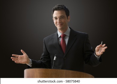 A businessman is standing at a podium with a microphone giving a lecture with outstretched hands. Horizontal shot.