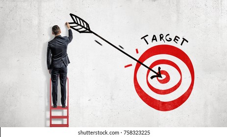 A Businessman Stands And Draws On The Wall A Big Arrow In The Center Of A Red Archery Target. Business And Success. Build Your Future. Reach Your Goals.