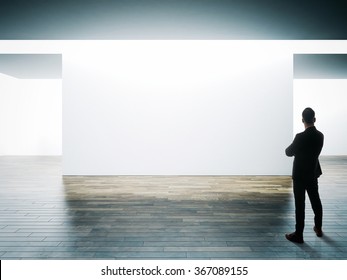 Businessman Stands Opposite Big White Wall In Museum Interior With Wooden Floor. Horizontal