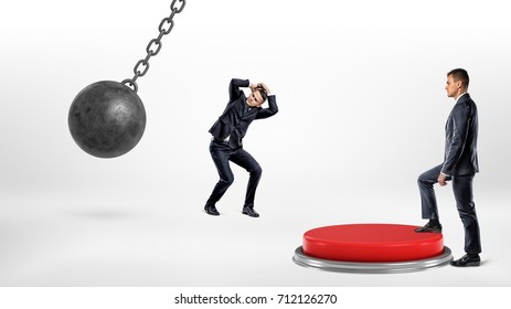A businessman steps up on a huge floor push button to protect another man hiding from a wrecking ball. Corporate world. Business competition. Help your colleagues.