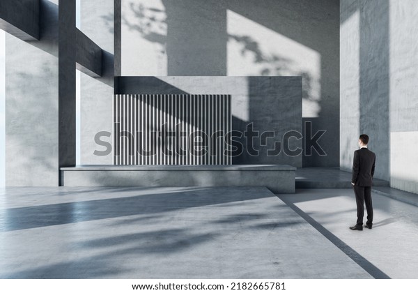Businessman in suit back view\
looking at blank black banner with place for your logo or text on\
dark grey wall in sunlit spacious hall with concrete design, mock\
up