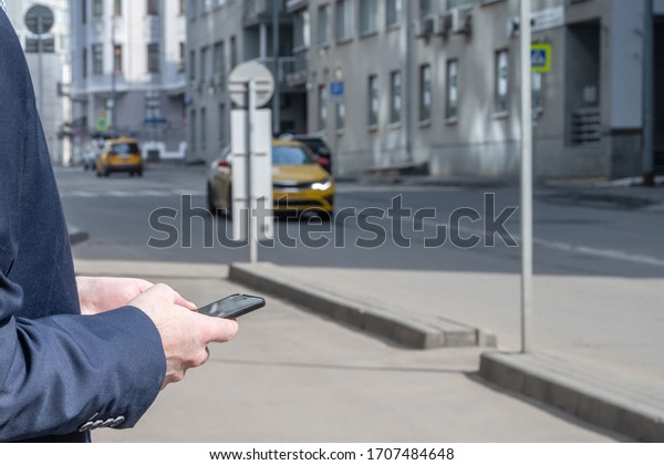 Businessman in a suit booking a taxi using mobile
phone app standing on the street. yellow taxi car on the background
of office buildings of the finance business center. Concept. Taxi
city service