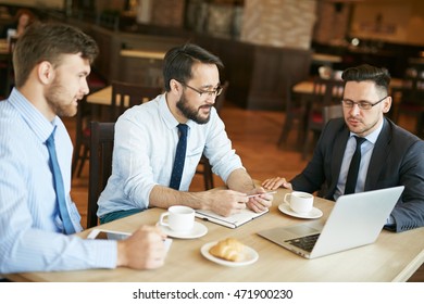 Businessman in suit and glasses working in cafe showing ideas on laptop to two pleased colleagues in shirts and ties with tablet and notepad on table